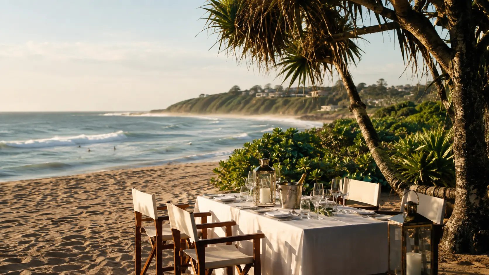 Private table set on the sand beneath a coastal palm as waves break in the late afternoon light